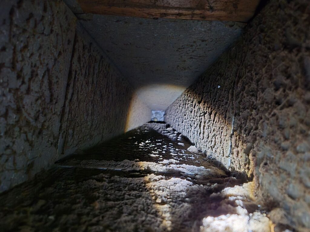 Inside of a dusty, rectangular air duct; light at the end, looking down the narrow, dirty space.
